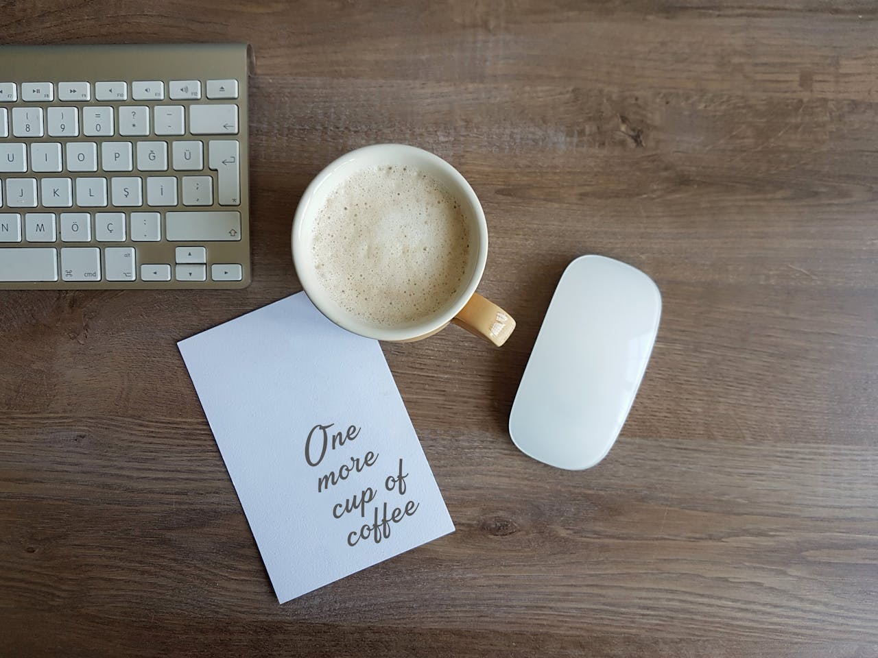 Top view of a workspace with a coffee cup, keyboard, mouse, and motivational card on wooden desk.