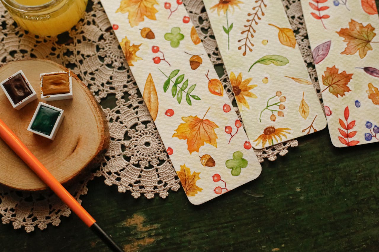 Colorful autumn-themed watercolor bookmarks on a crochet doily with painting supplies.